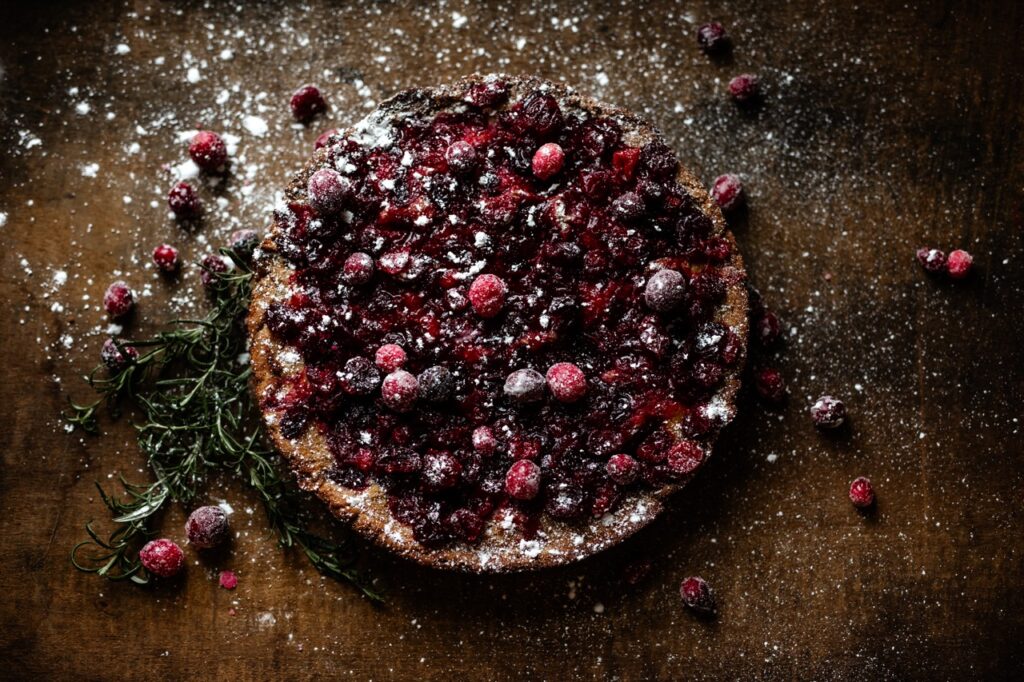 A spiced mascarpone cranberry cake with cranberries and powdered sugar on the table around it. 