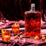 A glass filled with an amber colored negroni batch cocktail garnished with a candied ginger slice, illustrating the "Starlight Negroni" batch cocktail.