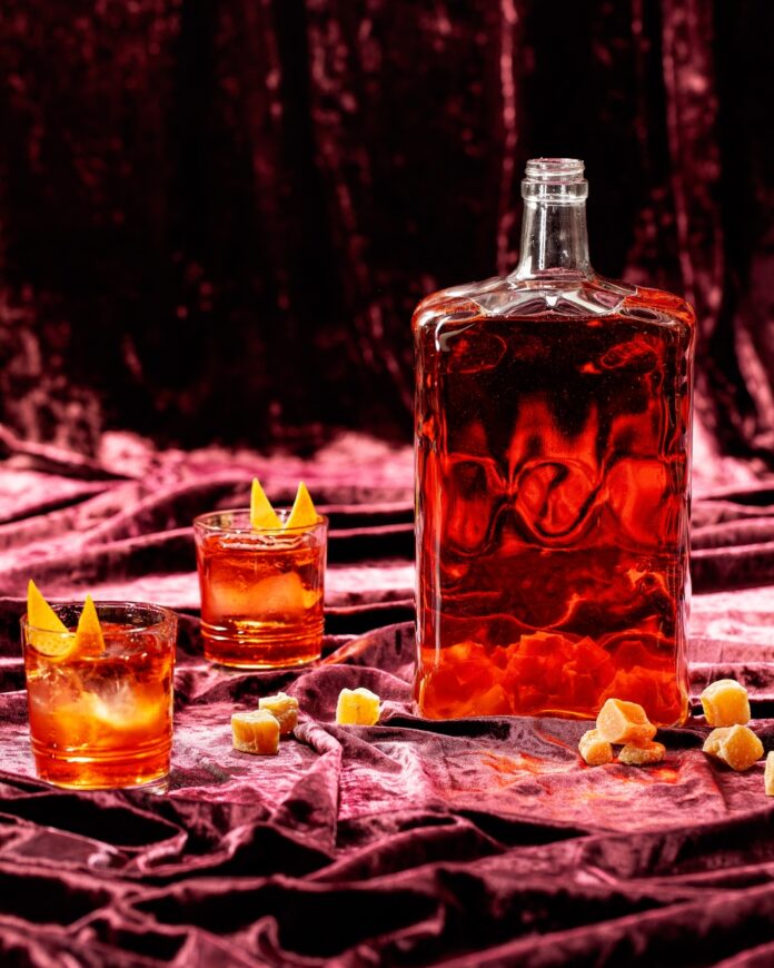 A glass filled with an amber colored negroni batch cocktail garnished with a candied ginger slice, illustrating the 