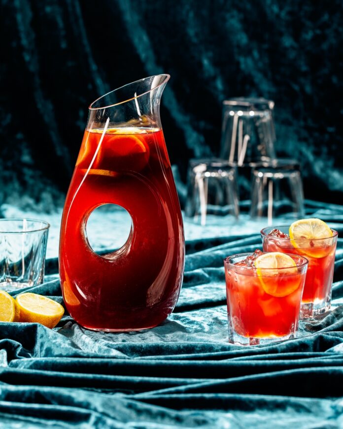 A pitcher filled with a deep red cocktail garnished with lemon slices, depicting the batched 