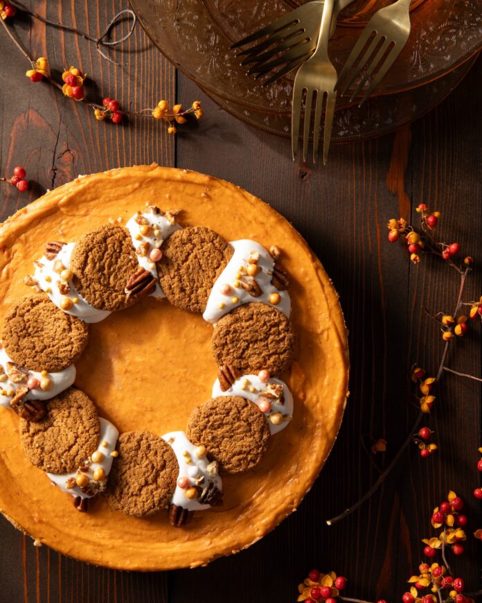 A Sweet Potato Cheesecake with a wreath of gingerbread cookies and whipped topping garnished with fall springs and pecans on a wooden surface.