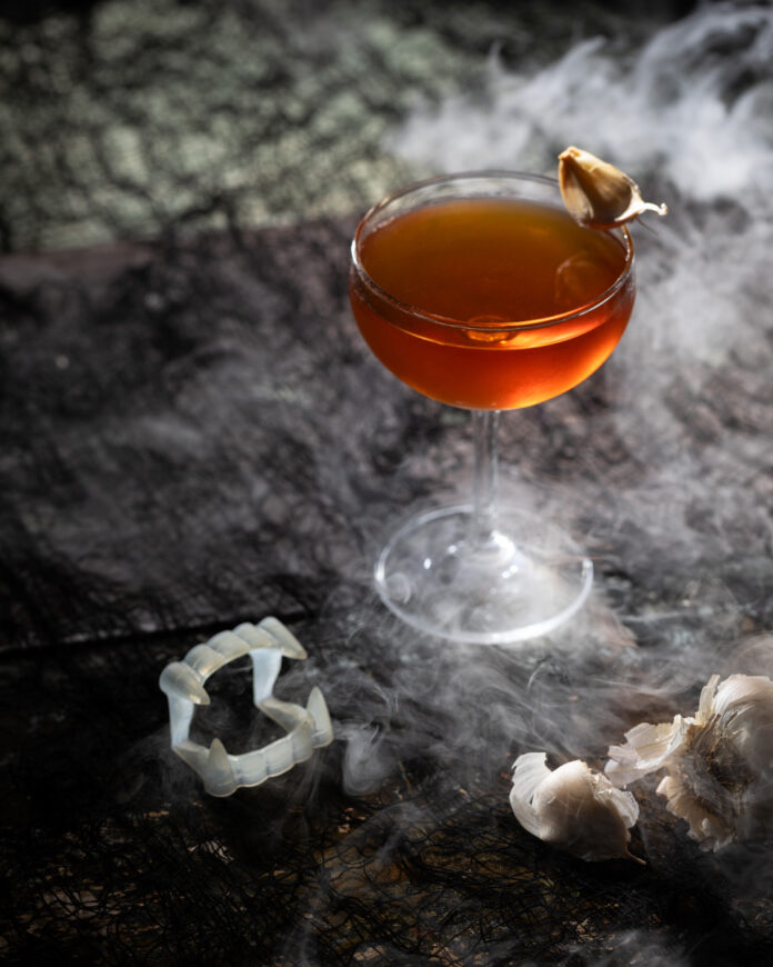 A bourbon cocktail with vampire teeth and garlic beside it