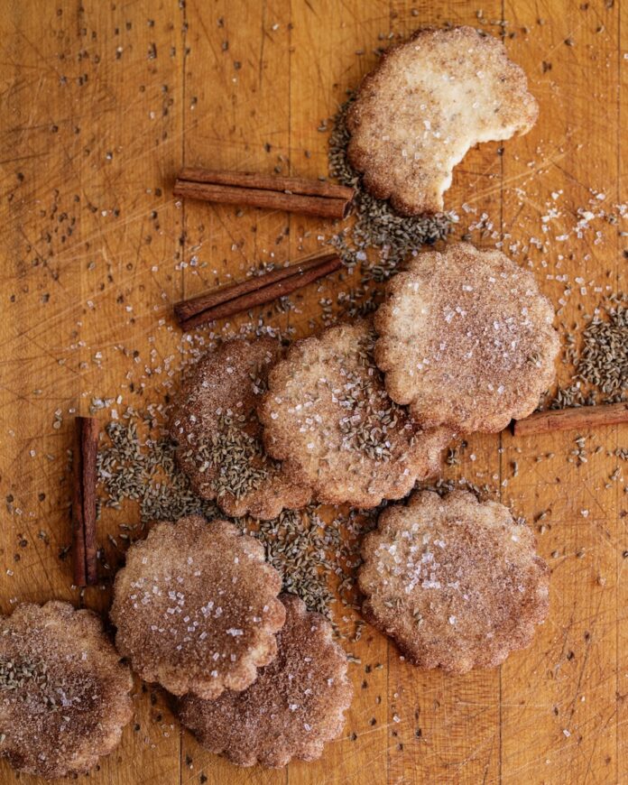 A group of traditional Biscochitos on a brown table surrounded by cinnamon sticks, sugar, and anise seeds.