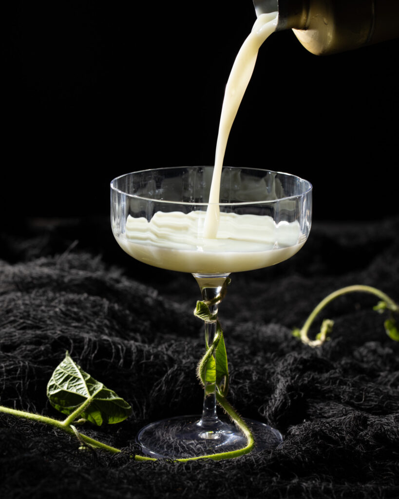 A person pours a white cocktail from a shaker into a coupe glass that's tangled in vines on a black background. 