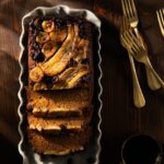 A Banana Ginger Upside Down Cake sits in a white loaf pan, cut into slices with bananas and raisins on top and two forks sitting beside the pan.
