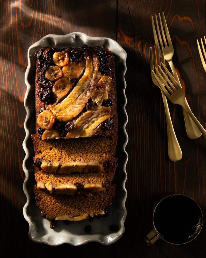A Banana Ginger Upside Down Cake sits in a white loaf pan, cut into slices with bananas and raisins on top and two forks sitting beside the pan.