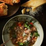 A bowl of brodo di carne soup with shredded beef, cannellini beans, and carrots, topped with fresh parsley and grated parmesan cheese, with bread and a wedge of parmesan on the side.