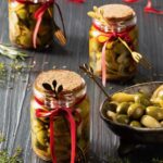 Three glass jars of marinated olives with cork tops and little gold forks tied to the jars with red ribbon sit on a black background with a bowl of olves to the right of them.
