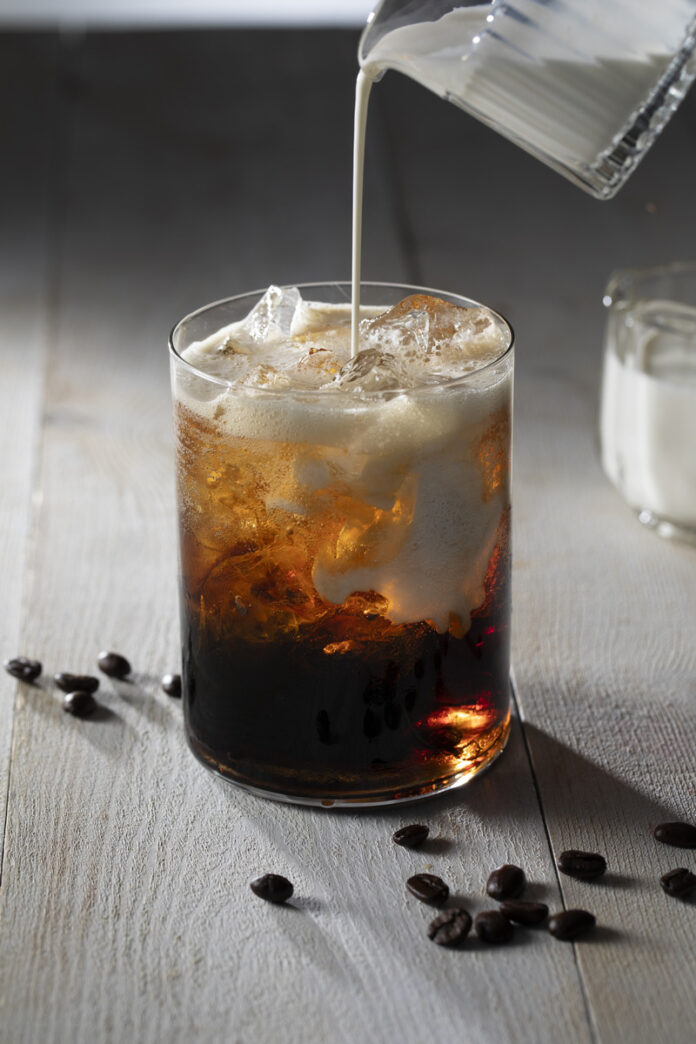 A glass filled with a dark, bubbly coconut cold brew dirty soda, with ice cubes and coffee beans scattered around it.