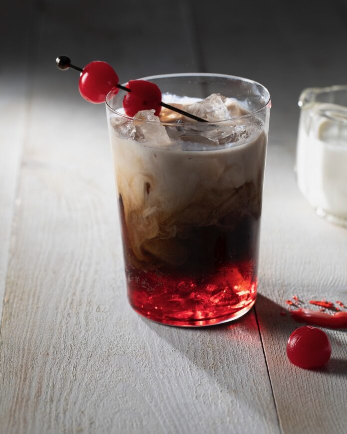 A glass of Dr Pepper with red syrup on the bottom, half and half on top, and a toothpick with cherries through it sitting on the glass.