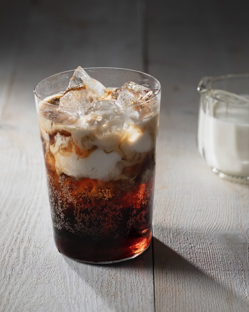 A clear glass holds Cherry Coke Dirty Soda with half and half and a small glass of half and half in the back right corner. 