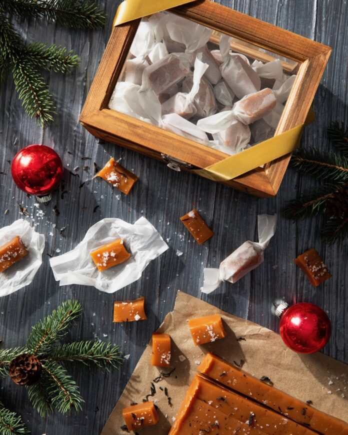 A glass box with wood edges holds wrapped caramels inside while unwrapped Ear Gray Salted Caramels sit below it on a black background beside a tee branch holding a red ornament.