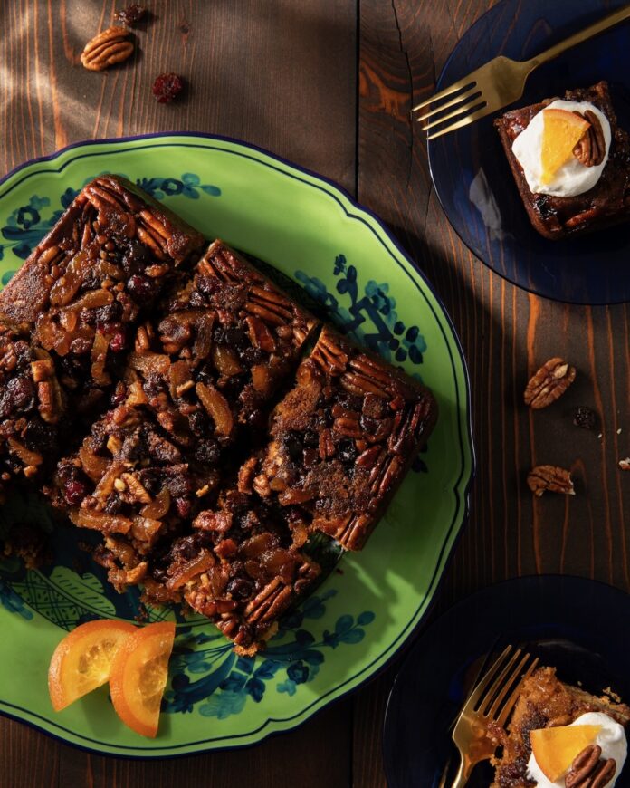 A square shaped upside down fruitcake cut into squares sits on a green plate surrounded by candied fruit slices and nuts.