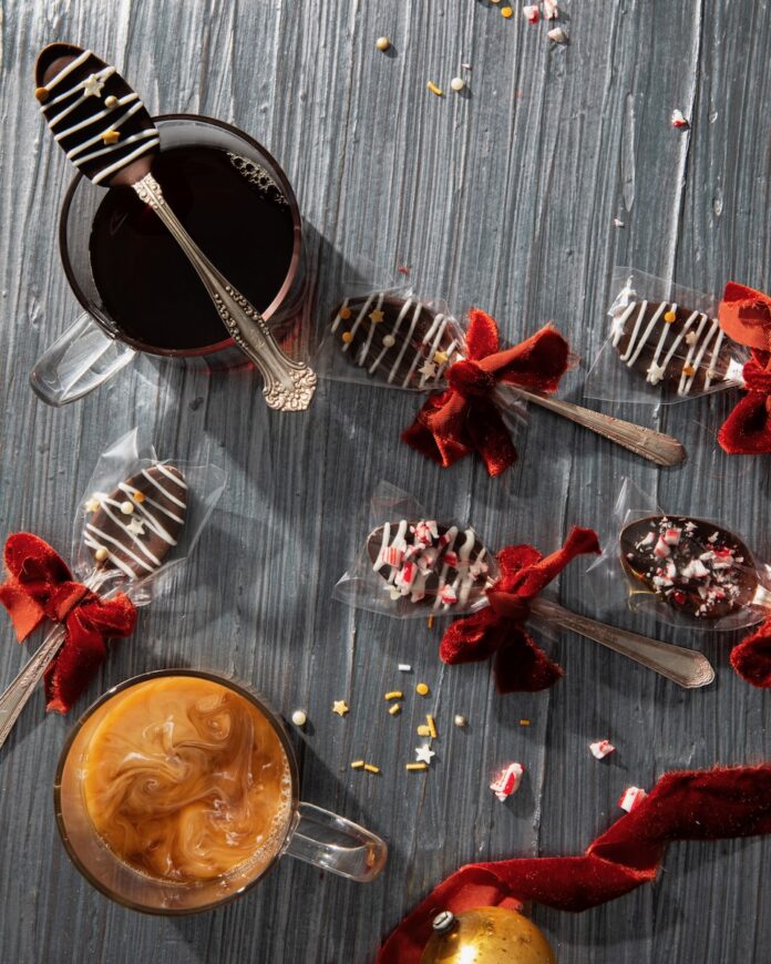 On a grey table sits various spoons coated in chocolate and toppings with a red bow around each as a cup of espresso and a latte sit to the left of the spoons.