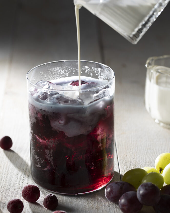 A glass filled with a deep purple-colored beverage, with ice cubes and what appears to be fresh grape slices floating in the liquid.