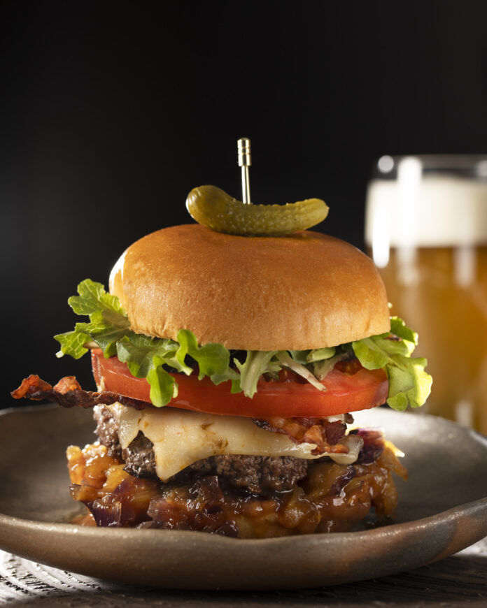 A delicious Hawaiian burger topped with bacon, pineapple, and red onion, served alongside a refreshing beer on a plate.