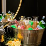 A metal bucket holds various bottled drinks along with carafes of non-alcoholic punch and a bowl of orange slices and cranberries sitting in front of the bucket.