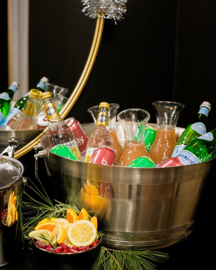 A metal bucket holds various bottled drinks along with carafes of non-alcoholic punch and a bowl of orange slices and cranberries sitting in front of the bucket.
