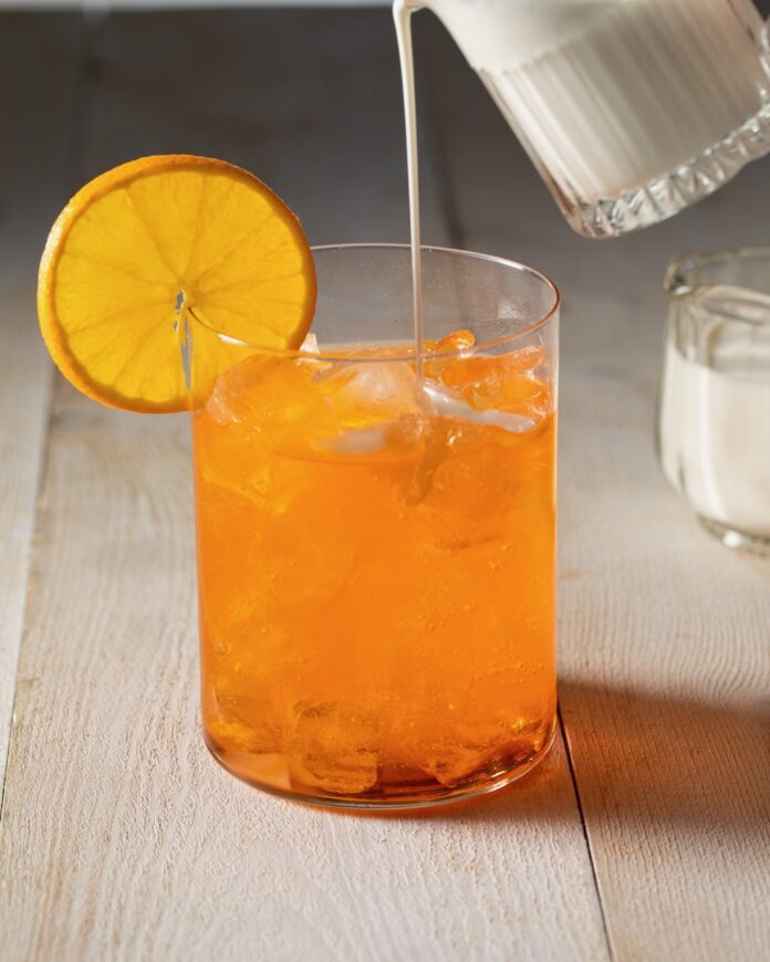 Orange soda sits in a short glass with an orange wheel on the rim as half and half is slowly poured into the drink to make a dirty soda.
