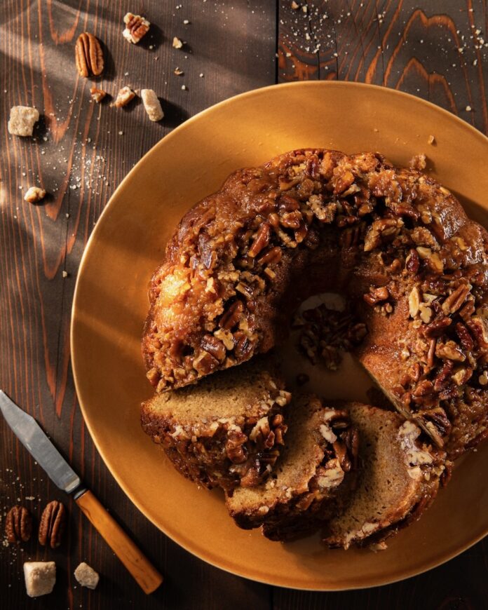 A pecan pie bundt cake half sliced sits on an orange plate with cropped nuts and brown sugar cubes around the plate.