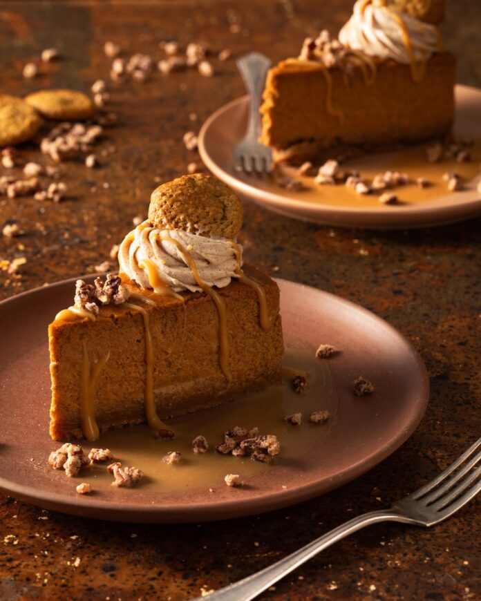 A slice of Pumpkin Cheesecake topped with whipped cream, nuts, a gingersnap cookie, and caramel drizzle sits on an orange plate with another slice on a plate in the top right corner and a fork in the bottom right corner.