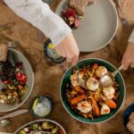 A shared meal of roasted carrots with crispy chickpeas and marinated labneh being served, set on plates with salad and bread on a patterned tablecloth.