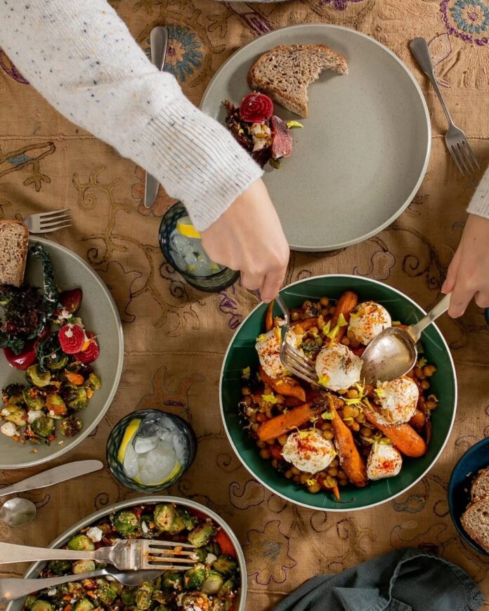 A shared meal of roasted carrots with crispy chickpeas and marinated labneh being served, set on plates with salad and bread on a patterned tablecloth.