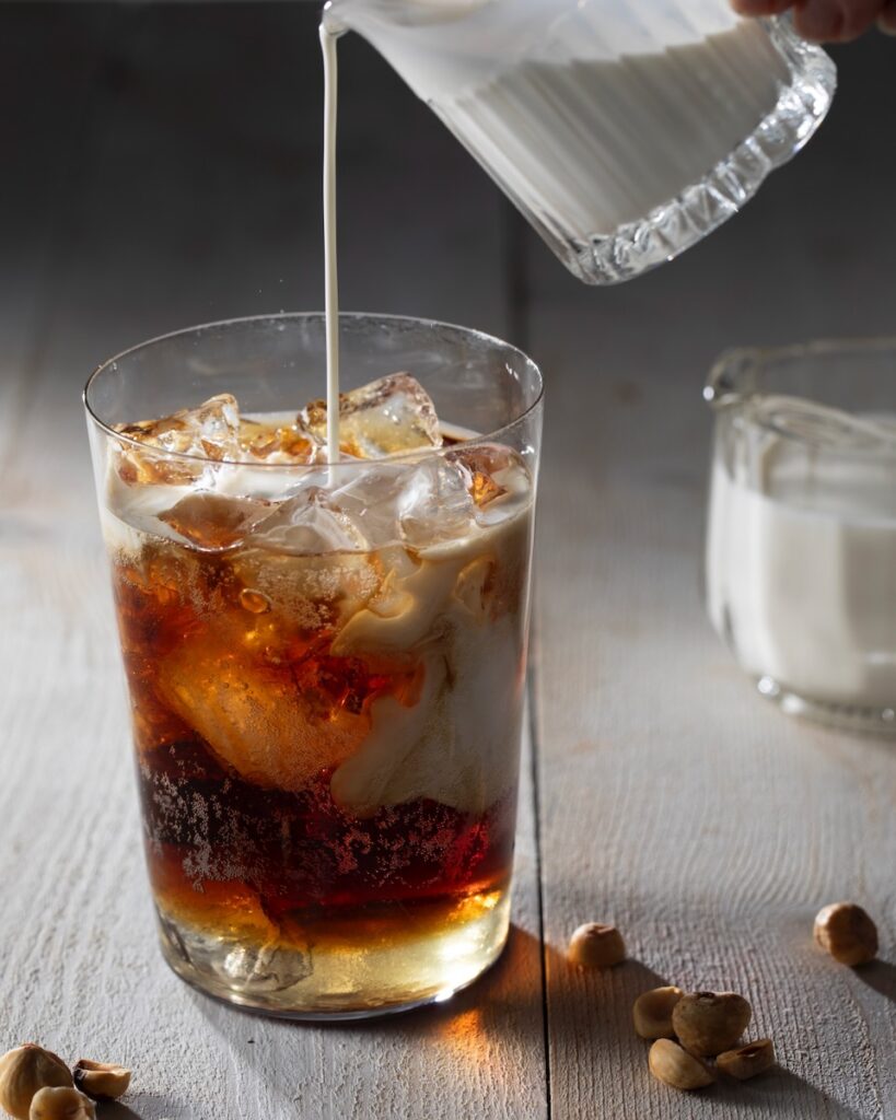 A person pours half and half into a cup of rootbeer to make a dirty soda with hazelnuts across the table and a small cup of half and half in the background. 