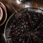 On a large metal platter sits a spiced cherry upside down cake cut into pie sized slices with a serving tool under one slice and powder sugar everywhere.
