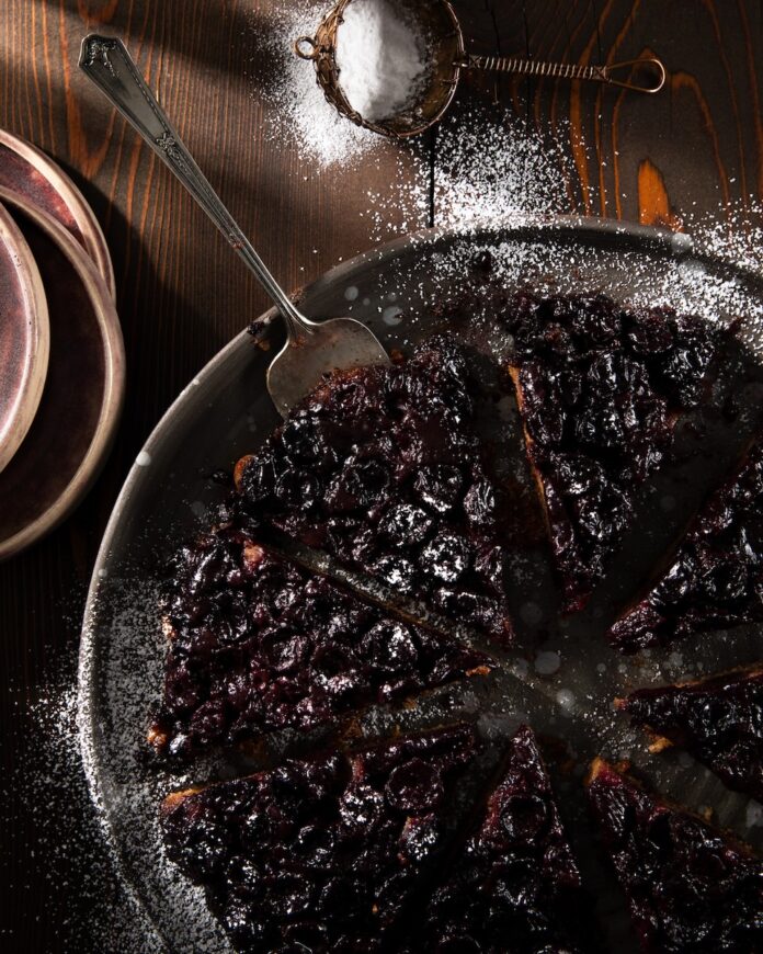 On a large metal platter sits a spiced cherry upside down cake cut into pie sized slices with a serving tool under one slice and powder sugar everywhere.
