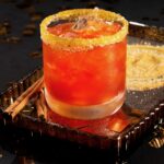 A brown sugar cider Manhattan, a red drink in a rocks glass rimmed with brown sugar and staged with cinnamon beside it