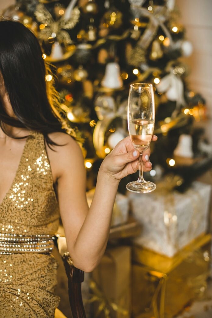 A woman stands in front of a Christmas tree holding a glass of sparkling white wine
