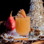 An orange Christmas mocktail staged with Christmas decorations and a red pear
