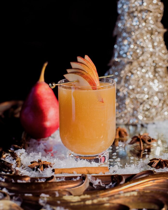 An orange Christmas mocktail staged with Christmas decorations and a red pear