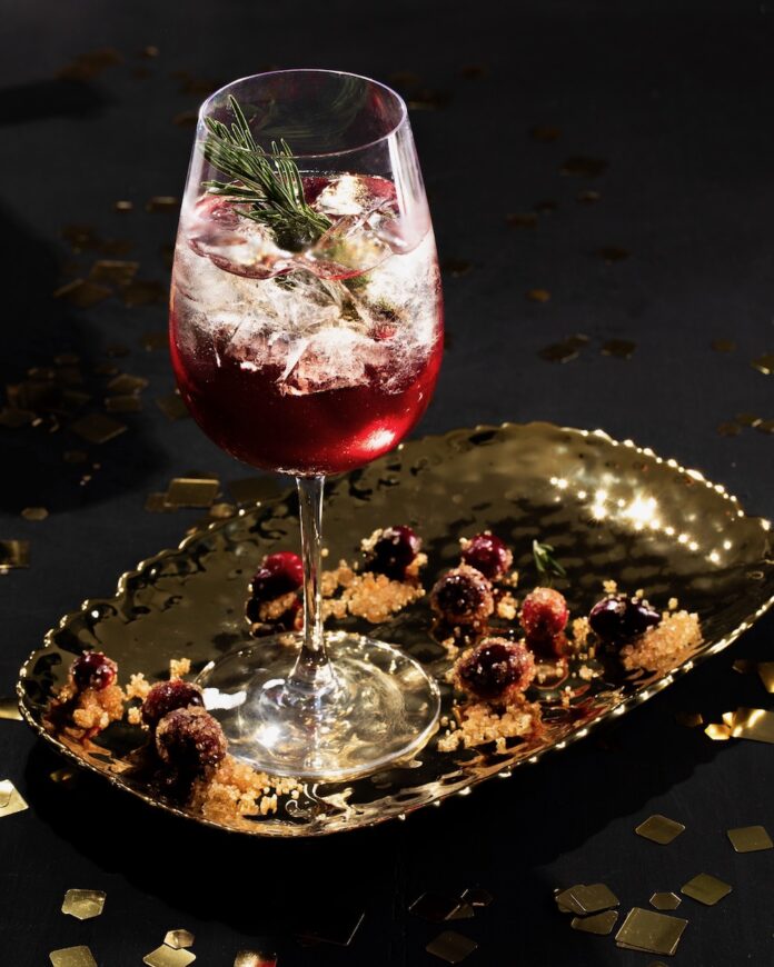 A rosemary cocktail in a white wine glass, gleaming with bright red cranberry juice and staged on a black background