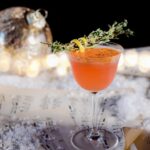 A spice cocktail staged over sheet music paper, garnished with a sprig of thyme and a lemon peel