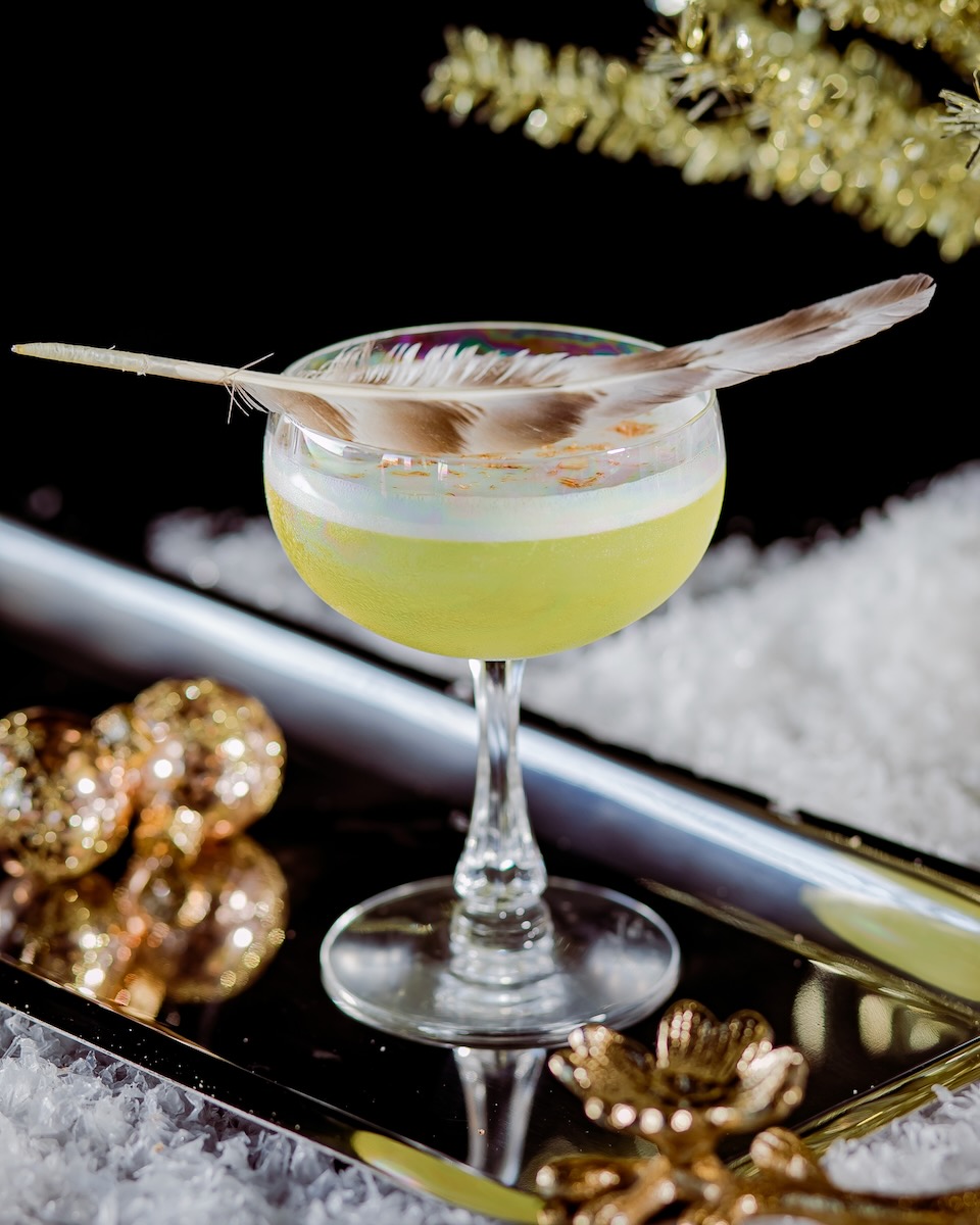A yellow French cocktail staged in a coupe glass and garnished with a feather