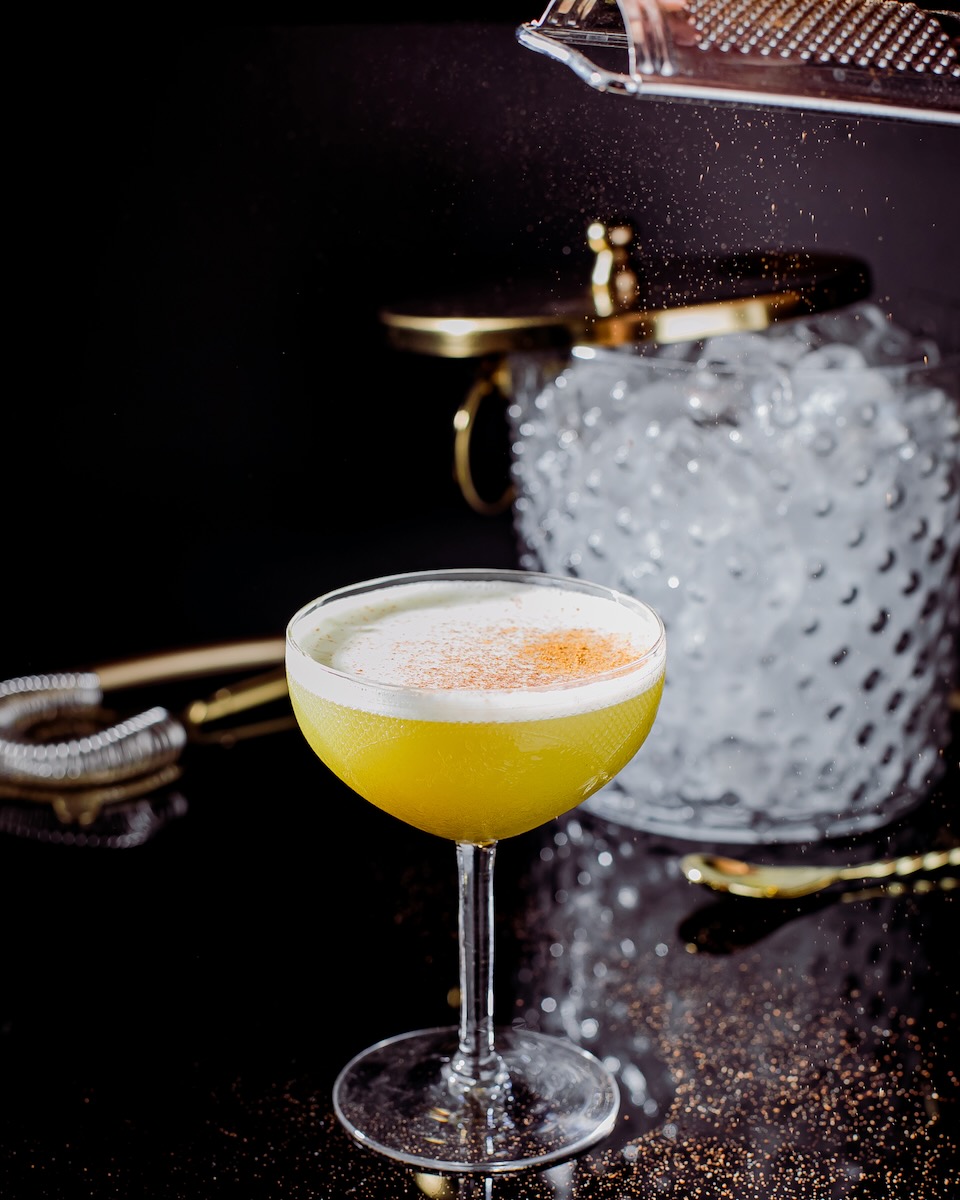 A yellow Grey Goose cocktail in a coupe glass, garnished with cinnamon and staged on a bar cart with several items behind it