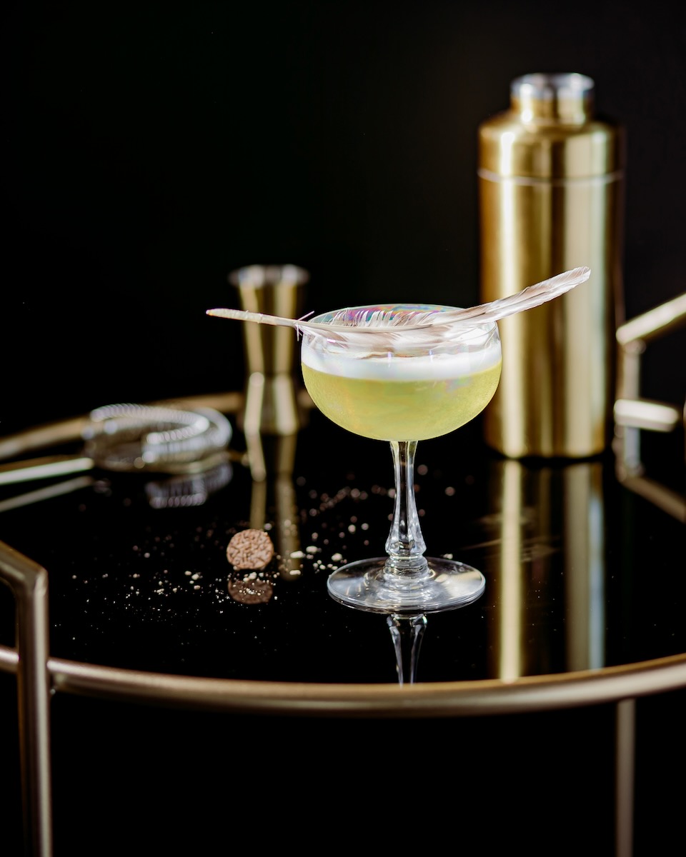 A yellow French cocktail garnished with a feather on a black background
