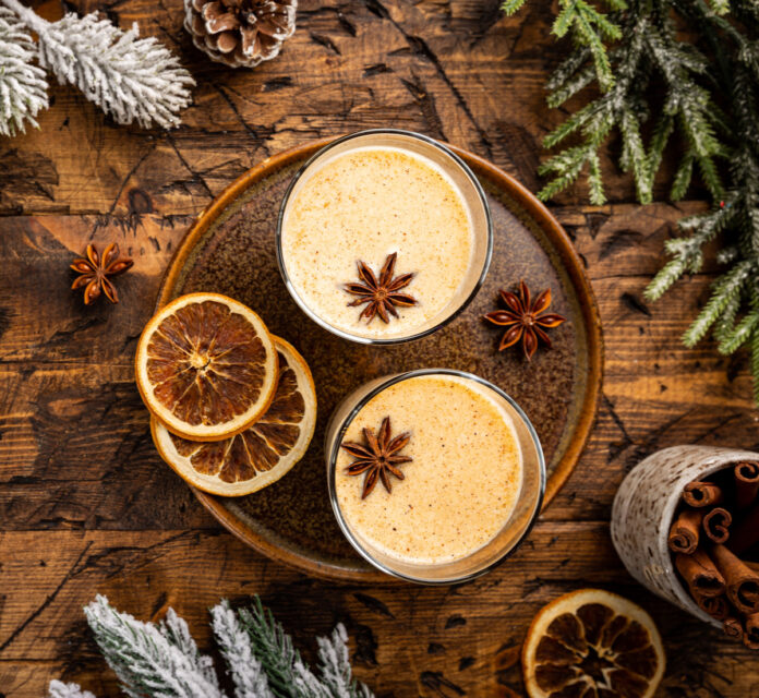 Two cups of eggnog garnished with cinnamon sticks sit on a brown plate