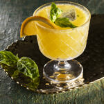 A glass of golden-colored cocktail with sliced orange and fresh basil leaves as garnish, set against a dark, mossy backdrop, creating a lush and earthy visual.