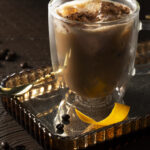 A glass mug filled with a dark, creamy-looking A non-alcoholic Irish coffee garnished with a lemon peel and what appears to be coffee beans or espresso grounds.