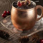 A copper mug filled with a dark, bubbly holiday mule non-alcoholic drink, garnished with sugared cranberries on a skewer.