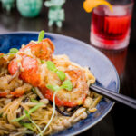 A visually appealing dish of long noodles with pieces of lobster, fresh scallions, and other vibrant ingredients arranged on a blue-gray plate.