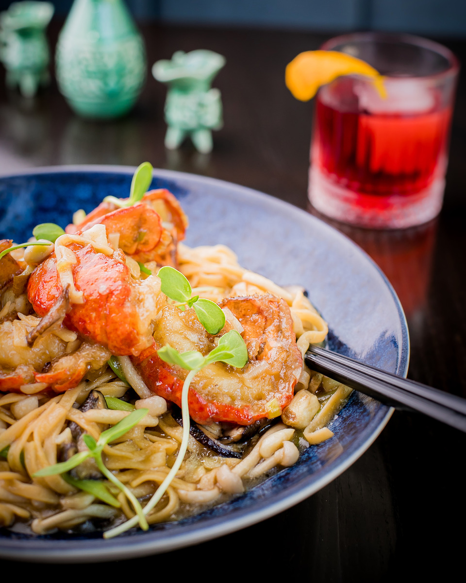 A visually appealing dish of long noodles with pieces of lobster, fresh scallions, and other vibrant ingredients arranged on a blue-gray plate.