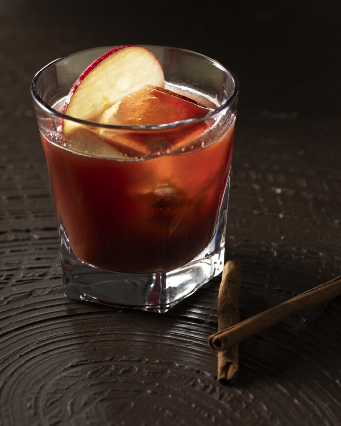 A rustic non-alcoholic old-fashioned cocktail served in a rocks glass, garnished with fresh apple slices, a cherry, and featuring a dense, foamy head.