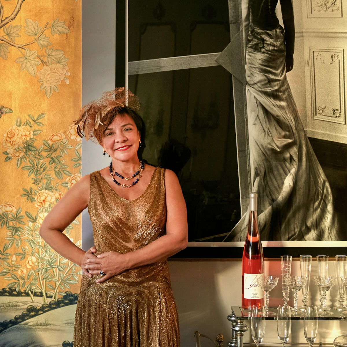 Maribel Lieberman stands beside a red, gleaming bottle outside a window, dressed in a golden dress