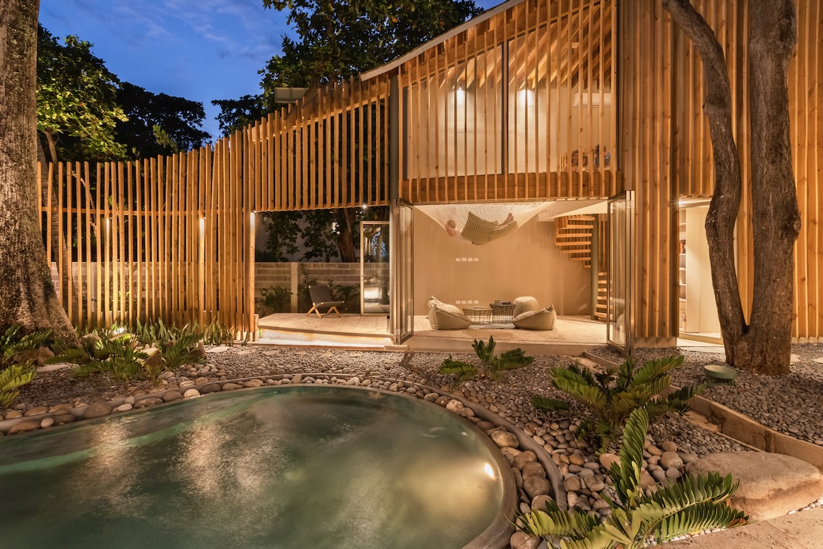 The exterior of Al Suave House at night, a rustic pool and wooden slat home 