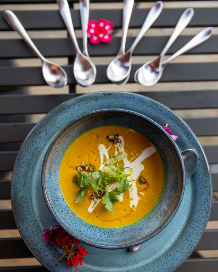 vibrant orange-colored soup served in a speckled blue-gray bowl, garnished with a dollop of coriander crème, crunchy pepita brittle, and fresh scallions and cilantro leaves.