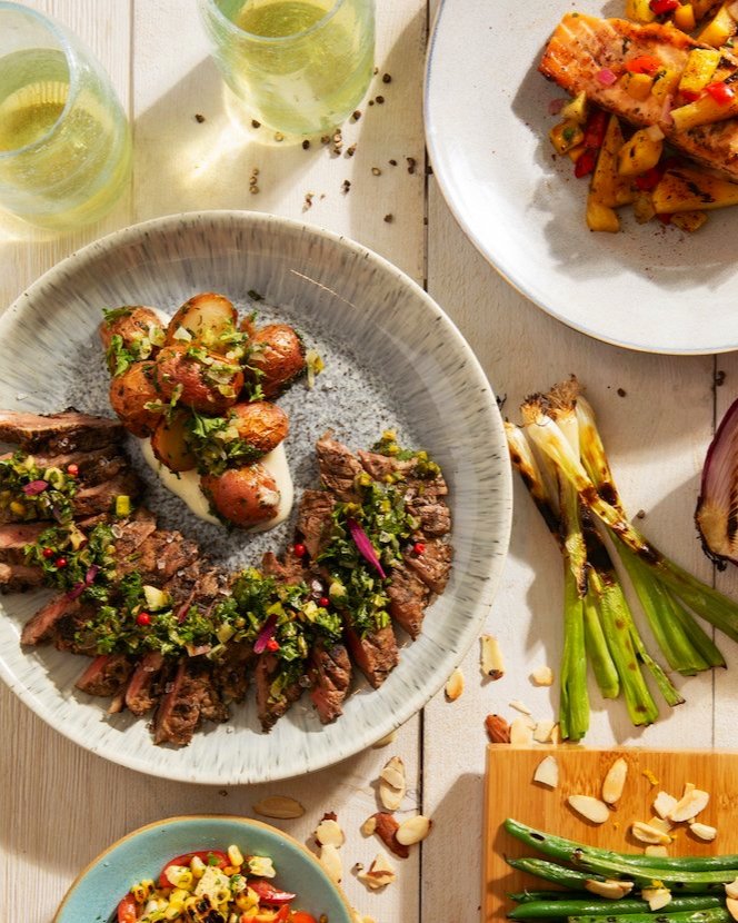 A white plate holds slices of grilled skirt steak topped with a scallion salsa as other summer dishes sit on the white picnic table nearby.